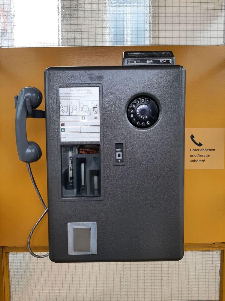 The image shows a vintage public payphone mounted on a yellow wall. The payphone is black with a textured surface and features a rotary dial with numbers 0 to 9, a handset on the left, and a coin slot at the top. The coin slot has labels for 